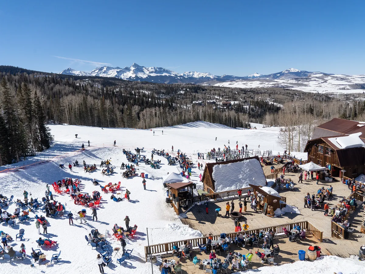 A lively scene at the base of the ski slopes, featuring people skiing and socializing near an outdoor seating area with a wooden lodge and mountain backdrop. The image is festive and bustling, capturing both activity and relaxation.