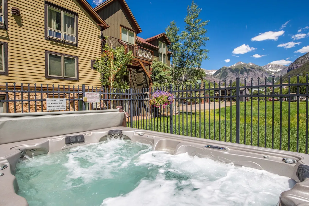 A serene exterior view of a hot tub with a fenced garden and mountain views in the background. The tranquil setting evokes the idea of soaking tired muscles after skiing or enjoying a quiet summer evening, with the property’s facade visible to connect the amenity to the home itself.