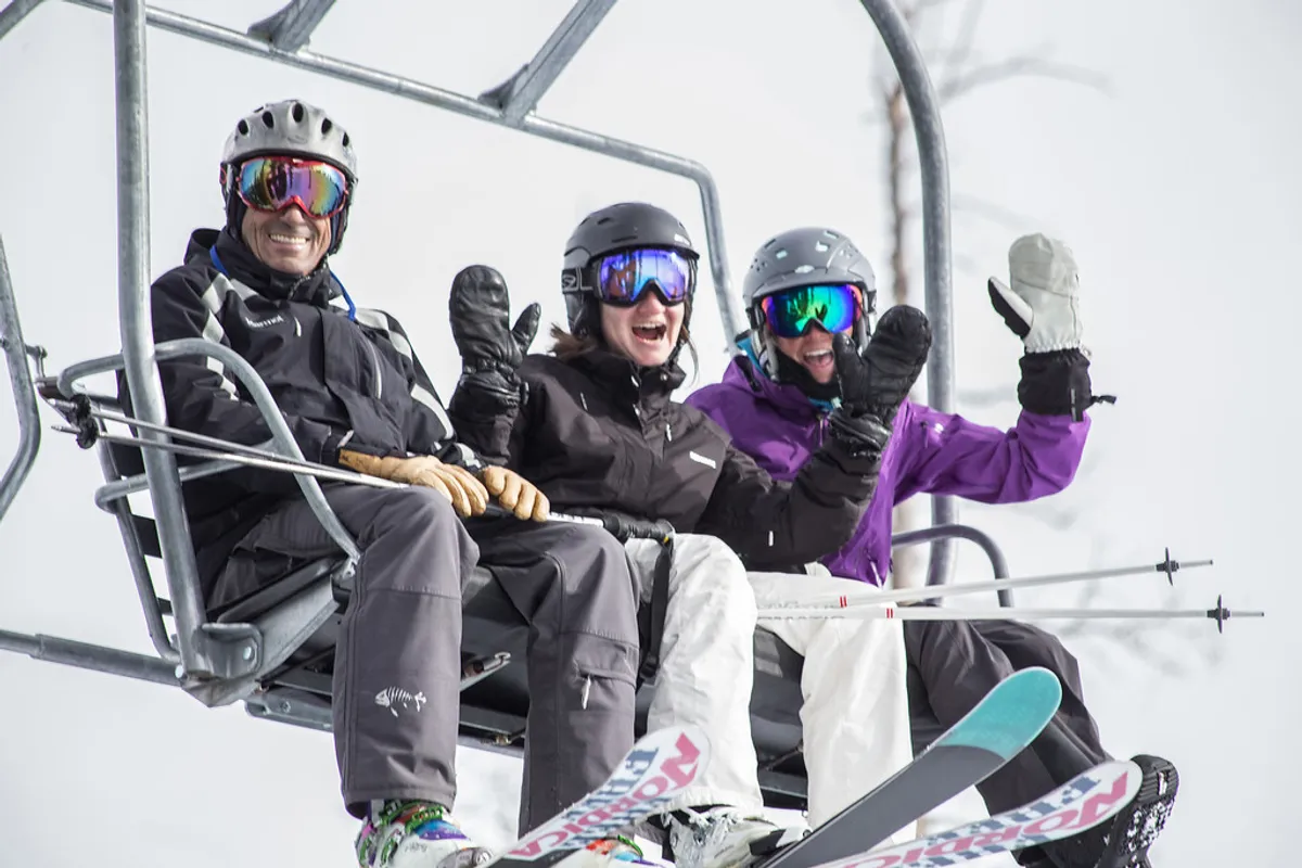This exterior image displays three skiers on a chairlift ascending a snowy mountain, with ski equipment visible and a sweeping view of white, snow-covered slopes and pine trees. The mood is lively and adventurous, evoking the communal spirit of ski trips.