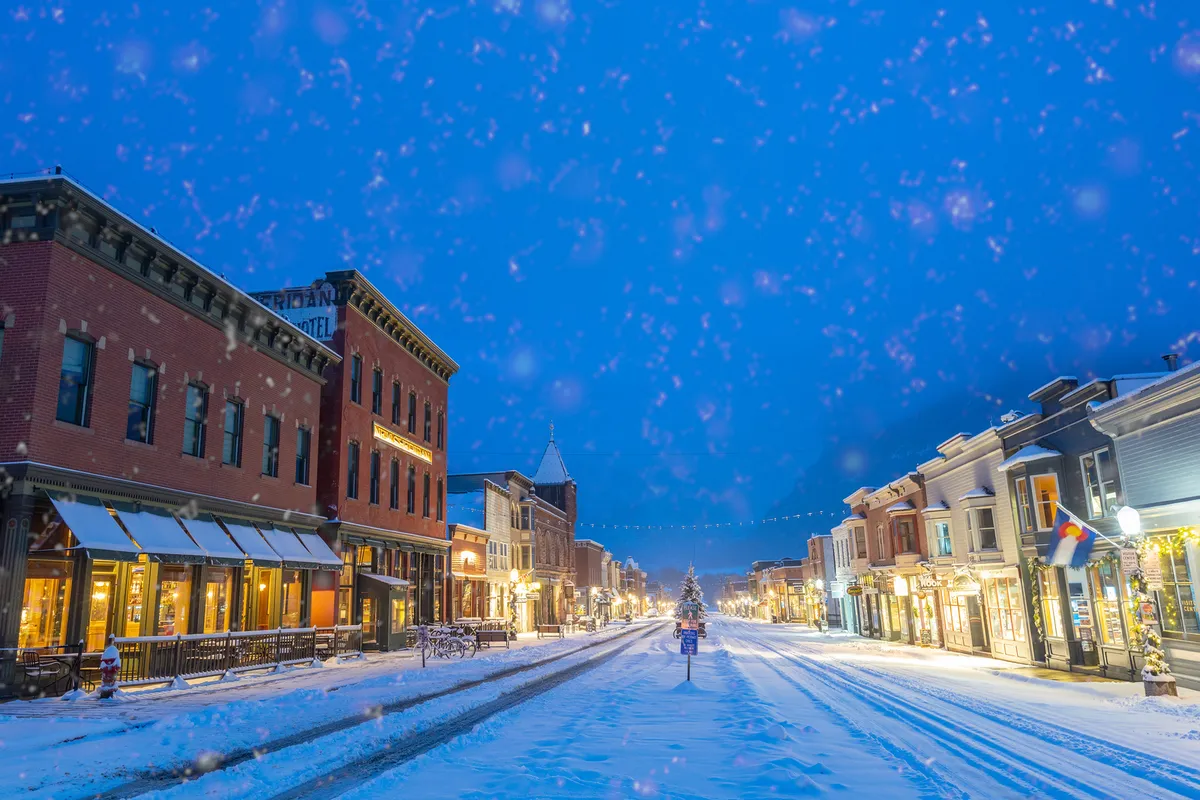 The festive image shows Telluride’s Main Street adorned with snow, twinkling decorative lights, a glowing Christmas tree, and historic storefronts. People can be imagined strolling, shopping, and enjoying the seasonal atmosphere, all within easy reach of the property.