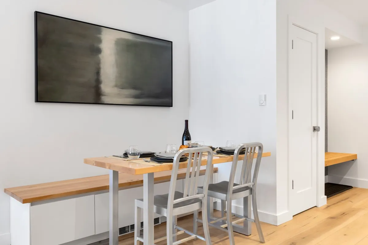 A modern dining area with a sleek table set for six, surrounded by contemporary chairs and highlighted by chic wall art. A bottle of wine sits on the table, setting the stage for intimate dinners and lively conversations. The room feels inviting, bright, and perfect for memorable group meals.