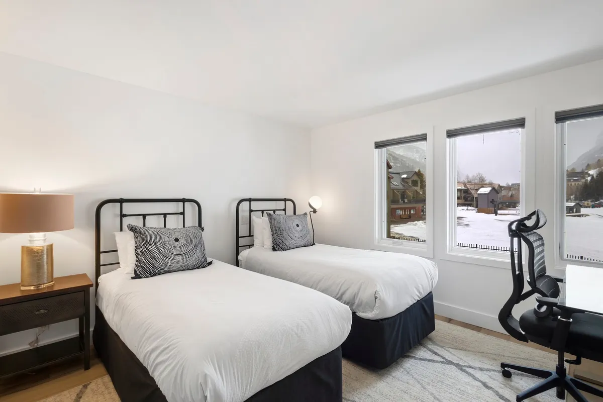 This image captures a bright bedroom with two twin beds, a desk and chair, and a lamp. The space is inviting and flexible, suitable for families, friends, or couples, and receives natural light, making it both restful and practical for quiet mornings or evening chats.