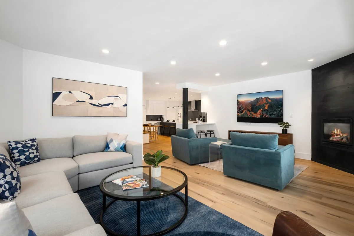 The image shows a spacious, sunlit living room with a large, inviting sectional sofa arranged around a modern glass coffee table. A cozy fireplace flickers beneath a mounted television, with contemporary artwork adding a stylish touch to the space. The room is designed for both comfort and socializing, perfect for relaxing, hosting game nights, or unwinding after a day on the slopes.