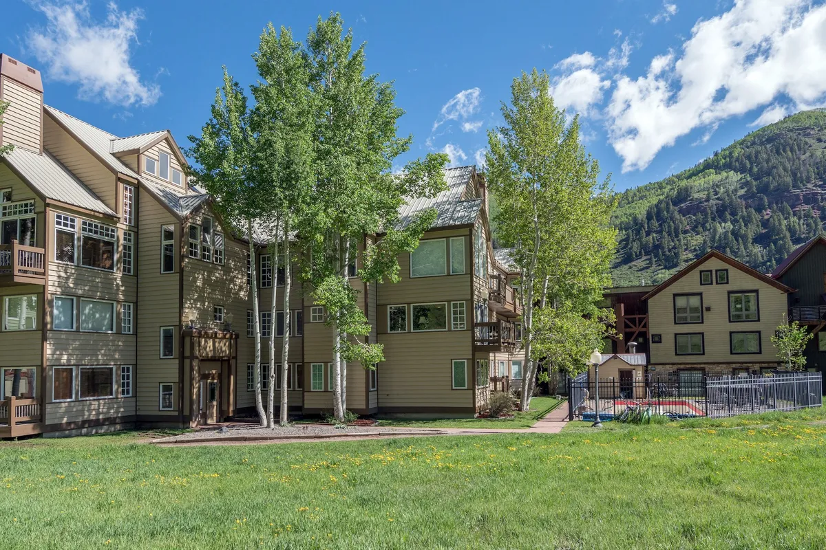 A welcoming multi-story building is nestled among trees and a landscaped lawn, with dramatic mountains as a backdrop. The exterior exudes warmth and hints at inviting communal spaces, perfect for gatherings both indoors and out.