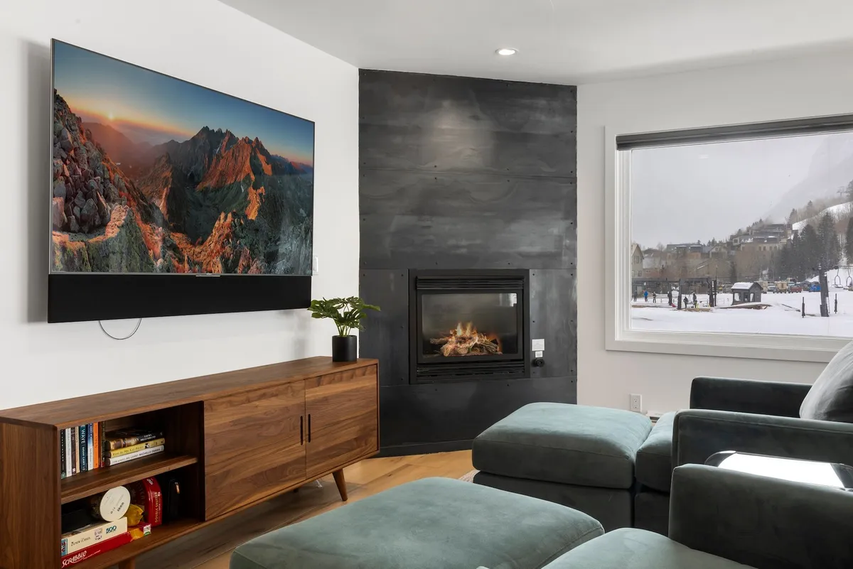 A cozy, contemporary living room featuring a modern fireplace, large television, and plush seating. Through expansive windows, you can see a snowy mountain landscape, providing a stunning natural backdrop to the inviting interior. The space is designed for relaxation and gathering, with the ambiance enhanced by warm lighting and stylish decor.