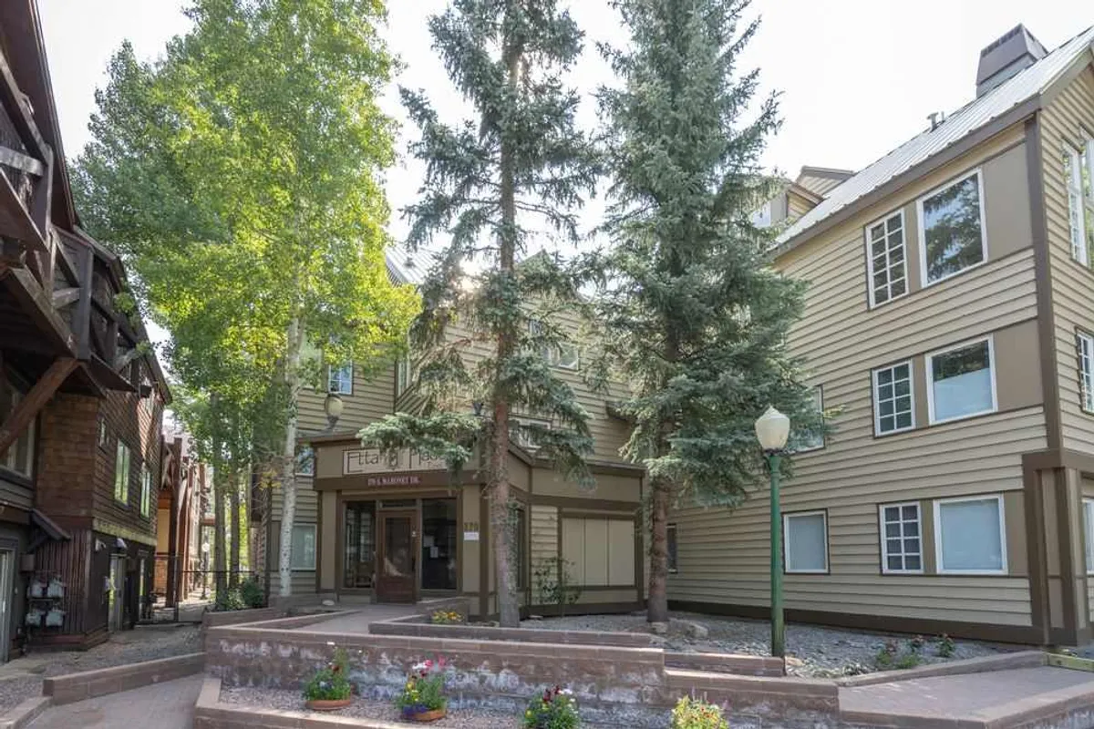 This exterior shot highlights the welcoming entrance to the property, framed by tall trees, colorful flower beds, and a well-maintained sidewalk. The scene suggests easy access to the surrounding neighborhood and a peaceful atmosphere just moments from Main Street’s bustle.