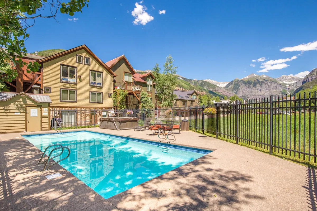 This image captures the inviting swimming pool surrounded by lounge chairs and set against a backdrop of majestic mountains and the building’s architecture. The sunlight highlights the pool area, emphasizing a leisurely, refreshing atmosphere ideal for summer relaxation.