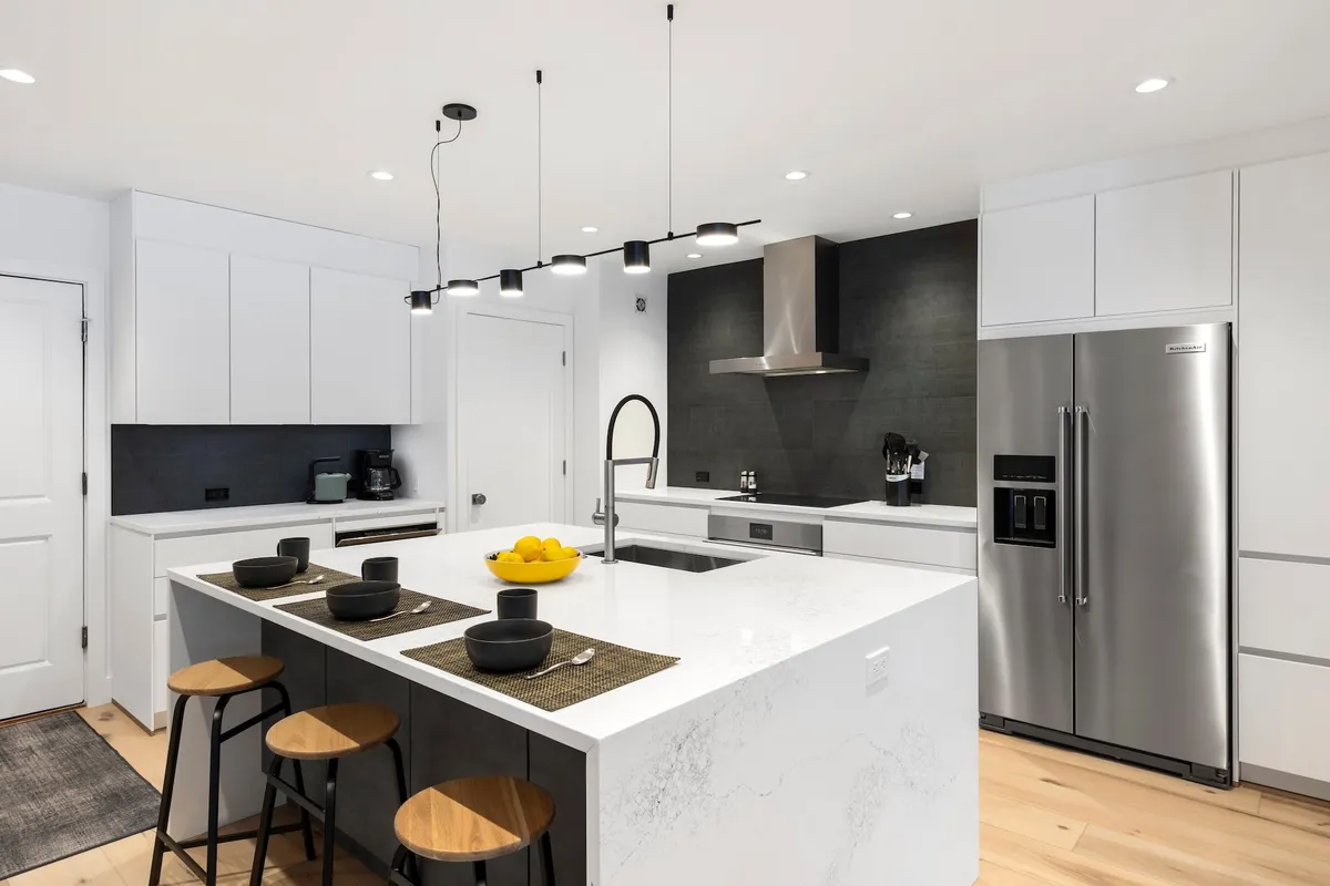 A large, modern kitchen with a central island featuring a marble countertop, surrounded by sleek stainless steel appliances. The color palette is black and white, creating a crisp and contemporary feel. The island is spacious, offering plenty of room for cooking and entertaining, and a wine fridge can be seen integrated into the cabinetry.